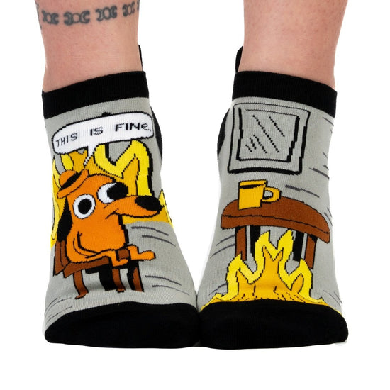 A model's feet wearing a pair of grey and back socks. One sock has a cartoon of a smilingbrown dog wearing a haat, sitting in a chair in front of burning flames. Above him is a caption saying "This is fine." The other sock has a brown wooden table with a coffee cup, and a roaring flame underneath it. The cuff and toes of the socks are black.