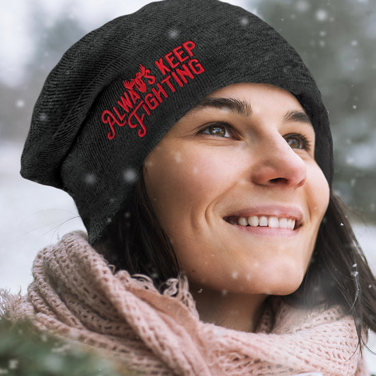 A female model in a snowfall, wearing a charcoal grey beanie. On the front is red text saying "always keep fighting." The "Y" in "always" is shaped like a moose head. Behind the model are snow-covered trees.