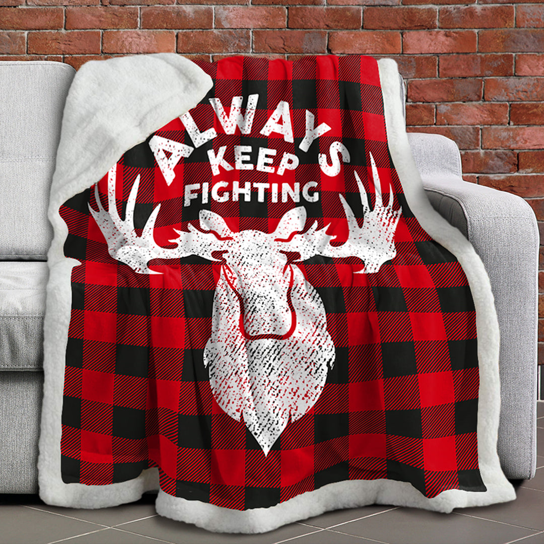 A red and black plaid blanket with a white border, slung over a white sofa. On the front of the blanket is a depiction of a moose head in white, with white text saying "Always Keep Fighting."