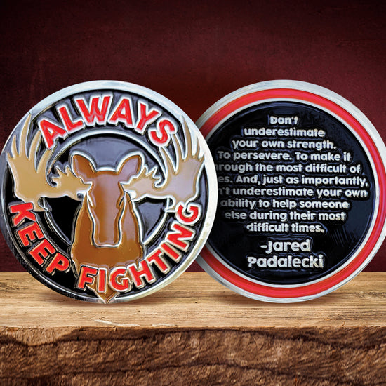 The front and back of a silver challenge coin. The front says "Always keep fighting" in bold red text, surrounding a brown moose silhouette. The back has a red circle around a black fill, in the fill it reads "Don't underestimate your own strength. To persevere. To make it through the most difficult of times. And, just as importantly, don't underestimate your own ability to help someone else during their most difficult times. -Jared Padalecki", in silver font.