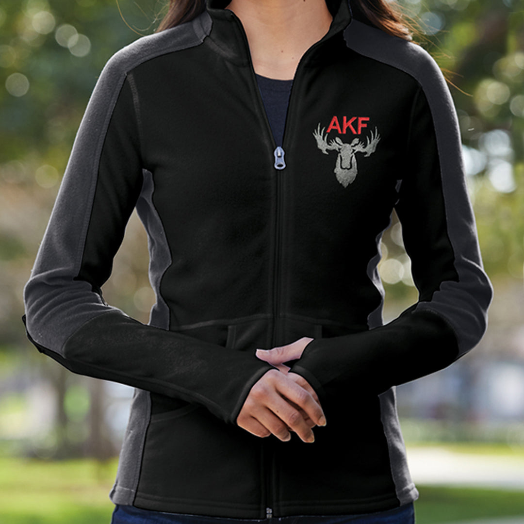 A female model wearing a black and gray fleece jacket. On the left lapel is red text saying AKF, above a gray moose head motiff. Behind the model is a field of trees..