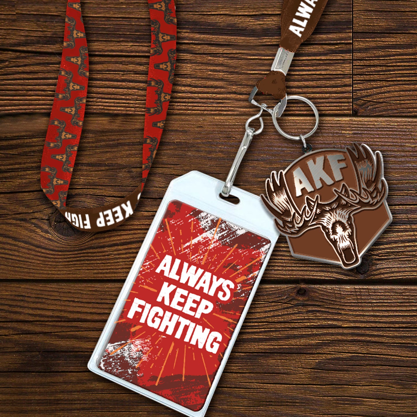 A red lanyard with white text saying "always keep fighting." Attached to the lanyard is a pendant depicting a moose head and antlers, with the letters AKF in silver at the top. At the end of the lanyard is a white tag with a red background and "always keep fighting" in white text. Behind the lanyard is a wooden table. In the bottom right corner is an inset image showing details of the strap, which has drawings of a moose head in a repeating pattern.