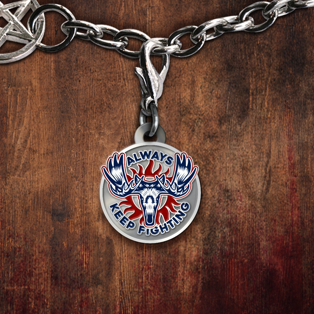 A silver-toned charm featuring a moose skull and the words 'ALWAYS KEEP FIGHTING' in blue, red, and white, displayed on a dark wood background.