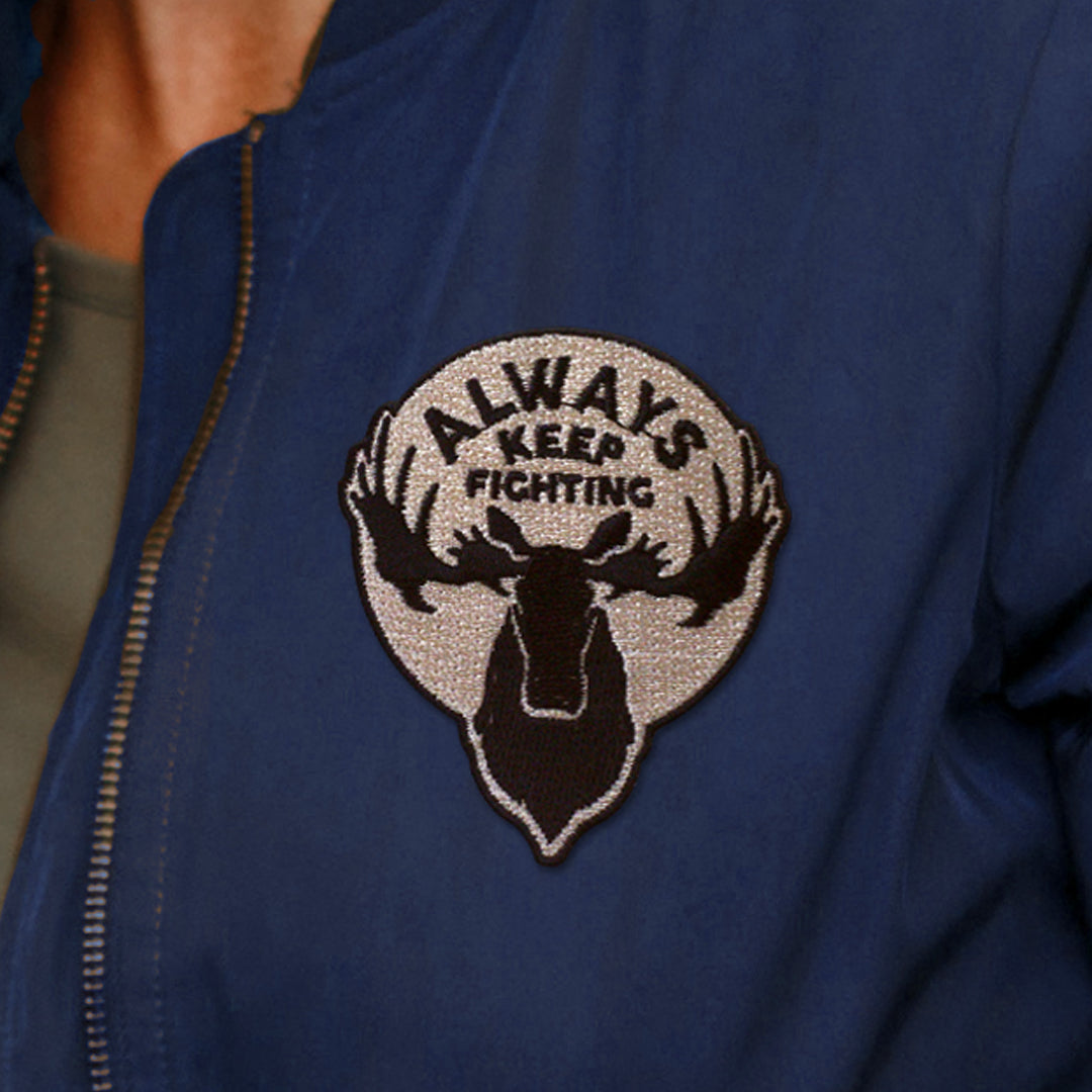 A tan, mostly circular patch with black edges, on a blue jacket. The words "Always Keep Fighting" are embroidered in black thread near the top, above a black embroidered moose head. 