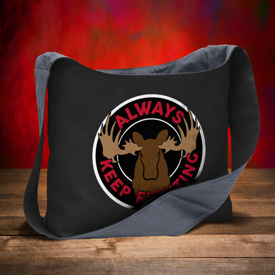 A black canvas sling bag on a wooden table, against a red background. On the front of the bag is a white circle with a brown moose drawing in the center. Around the edge is red text saying "always keep fighting."