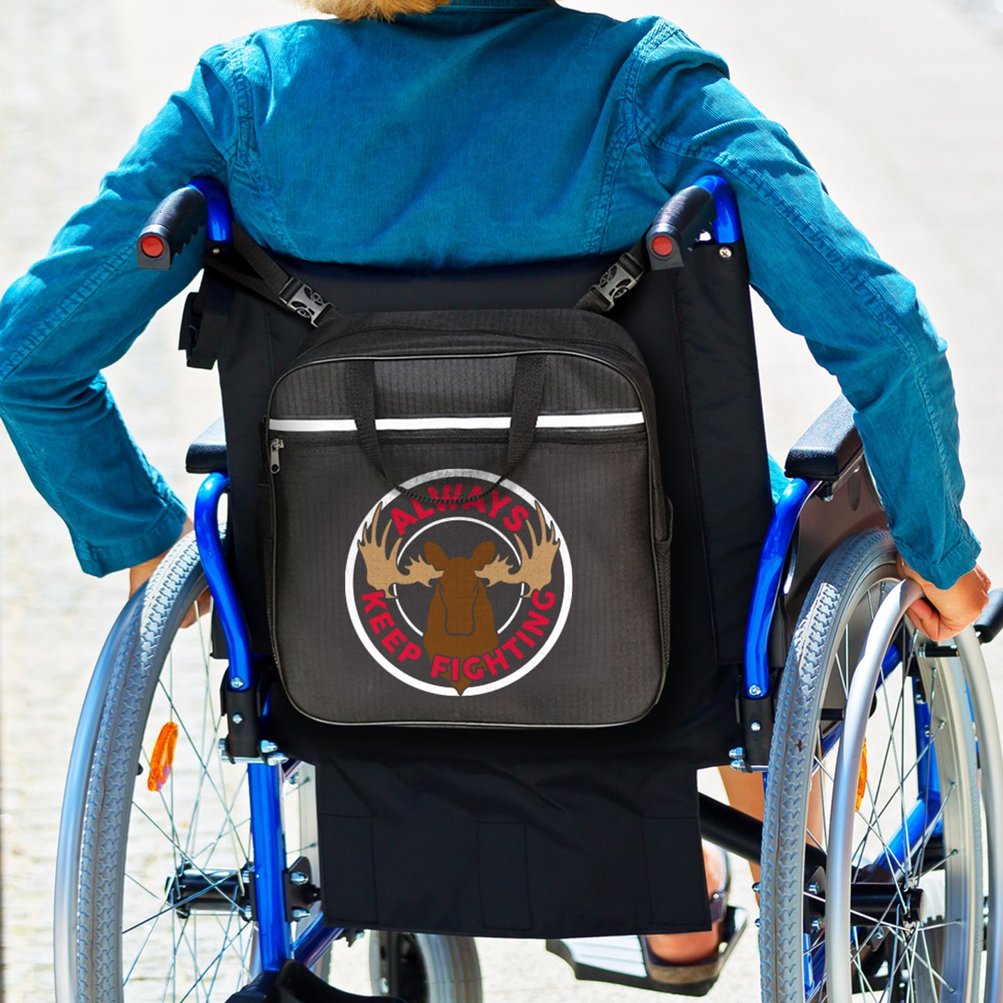 An image of a model in a wheelchair, rolling through a brick-lined ramp. On the back of the wheelchair is a black caddy bag, with a white circle around a drawing of a moose head. Red text around the moose says "Always Keep Fighting.