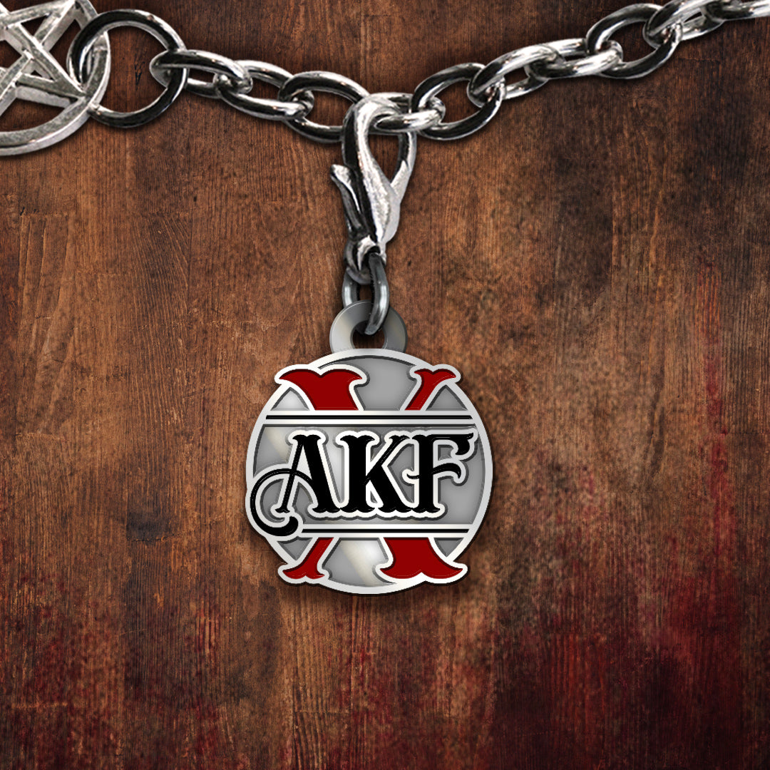 A silver-toned charm featuring the letters AKF in black over a red-and-silver crossed-design emblem, set against a dark wood background.