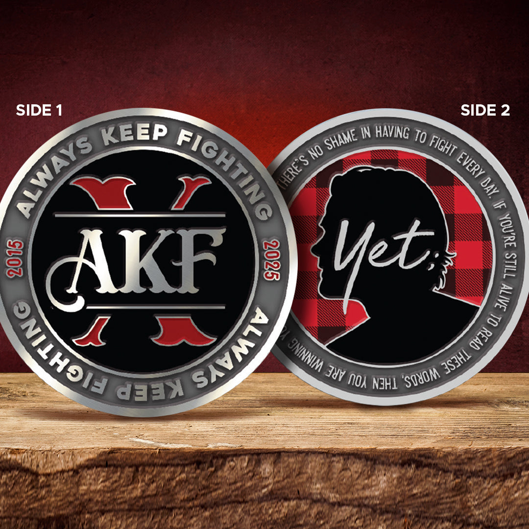 Two challenge coins side by side on a wood ledge. The left coin has "AKF" in white text above a red X on a black background. Raised text around the edge says "always keep fighting, 2025" The right coin has a person's head in silhouette, above a red and black plaid background. White text in the middle says Yet. Raised text around the edge says There's no shame in having to fight every day. if you're still alive to read these words, then you are winning your war, you're here.