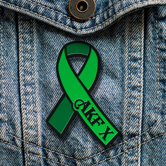 A green ribbon-shaped pin attached to a blue denim jacket. Black text on the ribbon says AKF X