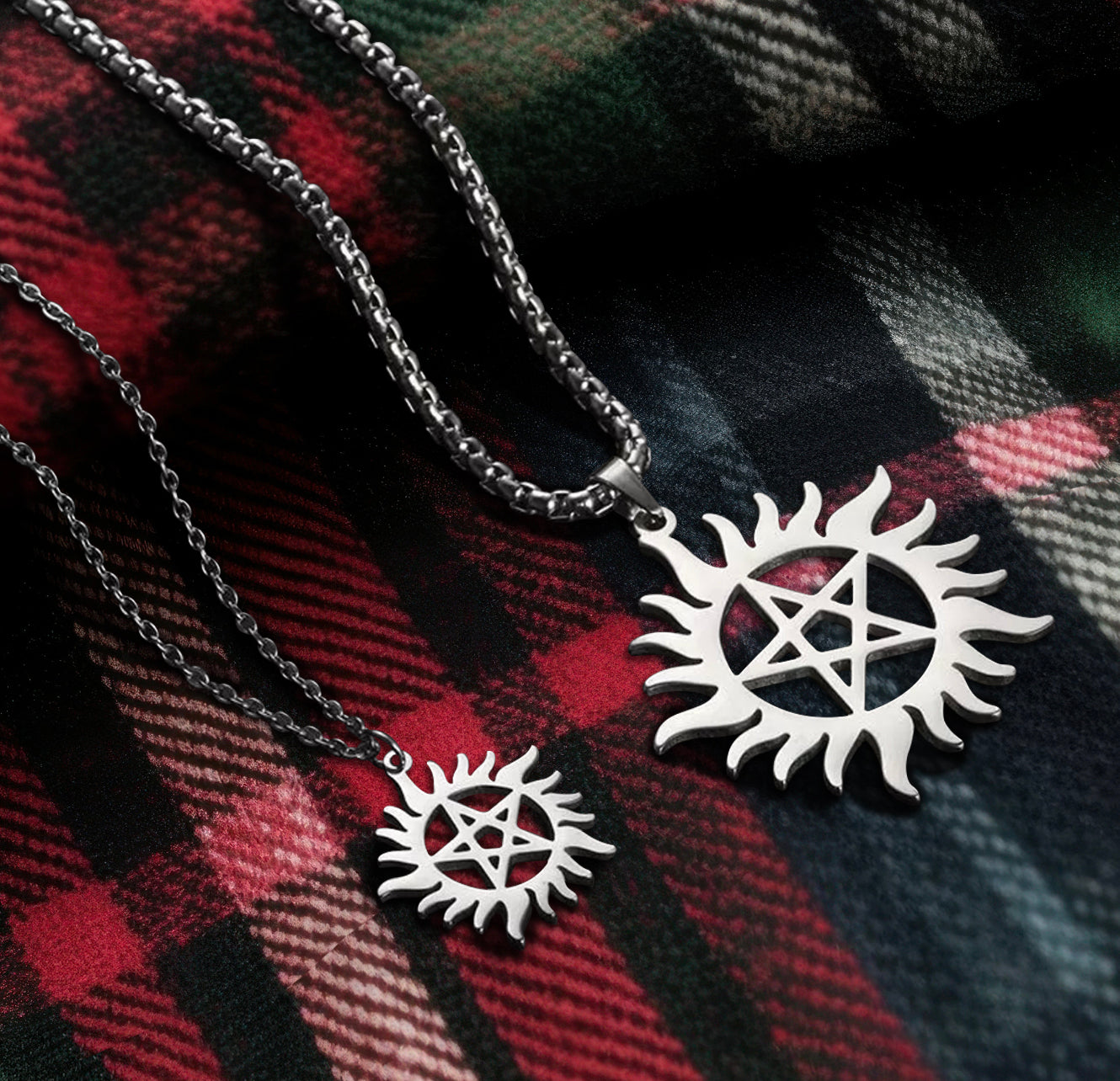 Two necklaces, side by side, on a red, blue, green and black flannel blanket. Both necklaces have pendants in the form of the Anti-Possession symbol. The pendant on the left is smaller than the pendant on the right. The left necklace has a link chain and the necklace on the right has a thicker, more sturdy chain.