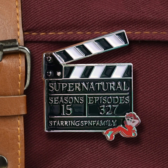 An enamel pin shaped like a film clapperboard, against a maroon messenger bag. Text on the front ays "Supernatural. Seasons, 15. Episodes, 327. Starring SPN family." At the bottom right corner is a female devil in red.