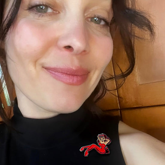 An image of actor Alaina Huffman. On her lapel is an enamel pin in the shape of a pin-up style woman lying back with her arms behind her head. She is wearing a red jumpsuit with devil horns and a devil's tail.