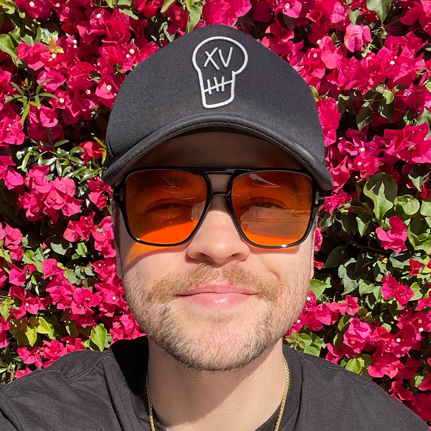 An image of actor Alex Calvert standing in front of a hedge covered in red flowers. Alex is wearing a black cat on his head. On the front of the cap is a stitching of a stylized skull, with the characters XV in place of the eyes.