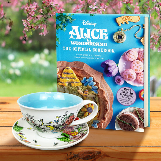 A ceramic teacup and saucer on a wood table, next to a cookbook. The teacup features drawings from Alice in wonderland. On the book cover is white text saying "Alice in wonderland, the official cookbook." Various cakes and cookies are shown on the cover, next to flowers and a pocketwatch. Behind the book and teacup is a field of grass and flowers.