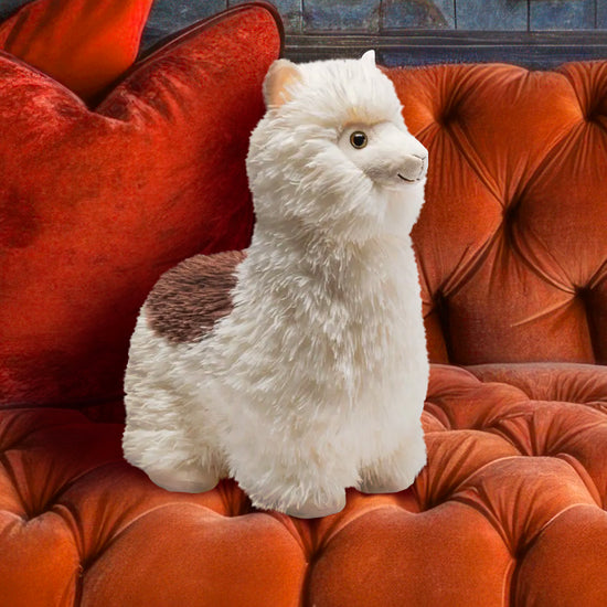 Close up view of a stuffed, smiling alpaca "sitting" on an orange-colored velvet couch. The alpaca is white, with a patch of brown fur on its back.