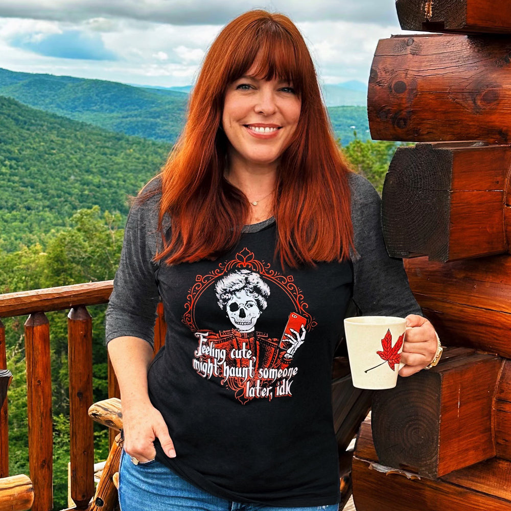 An image of Amy Bruni wearing a grey and black baseball shirt. She is standing on a wooden balcony overlooking a forest. On her shirt is a skeletal figure holding a red cell phone, surrounded by a red filigreed border. Under the figure in white text is "feeling cute, might haunt someone later, IDK."