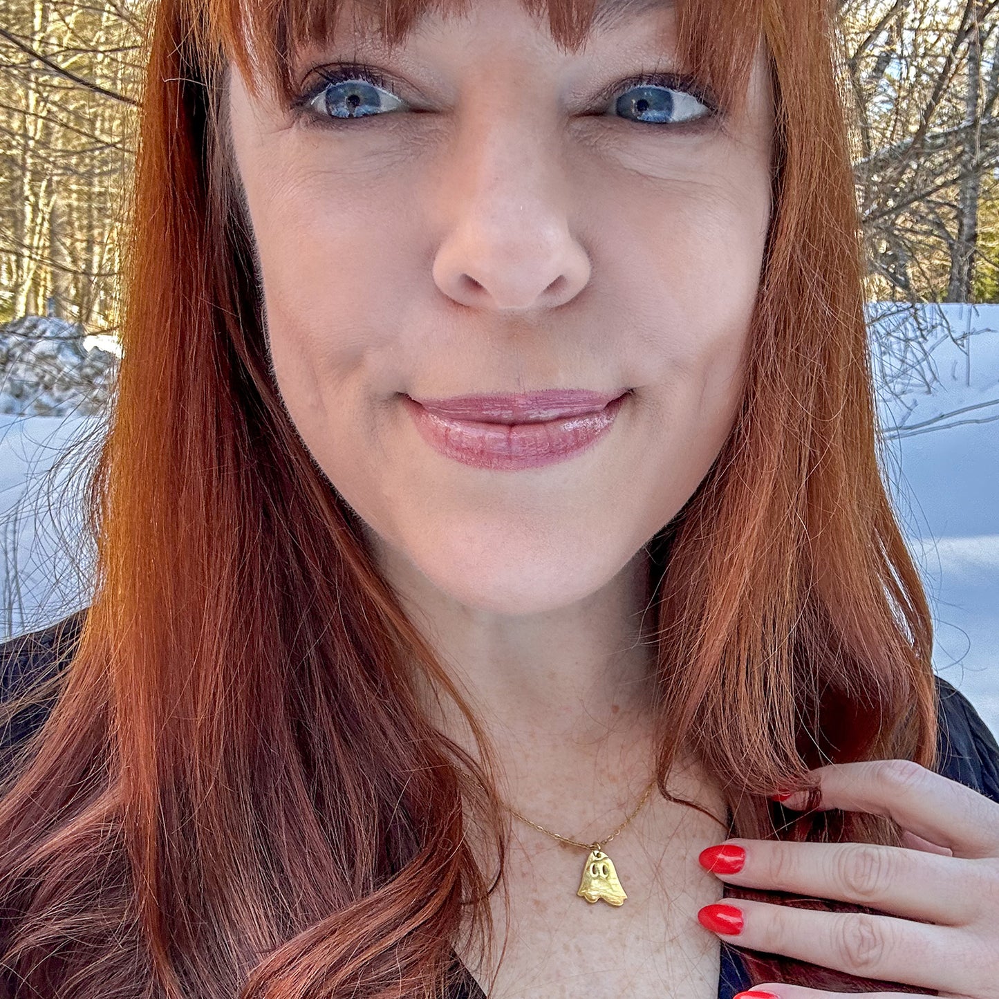 An image of paranormal investigator Amy Bruni, against a snow-covered field of trees. Around her neck is a small gold pendant in the shape of a ghost, with two spots cut out at its eyes. The necklace hangs on a gold chain.