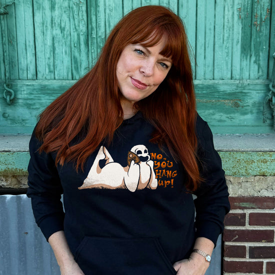 Amy Bruni poses in a black pullover hoodie. The print is a white ghost laying on its stomach with its feet up and a planchette pressed to its ear like a phone. Orange text reads "No, you hang up"