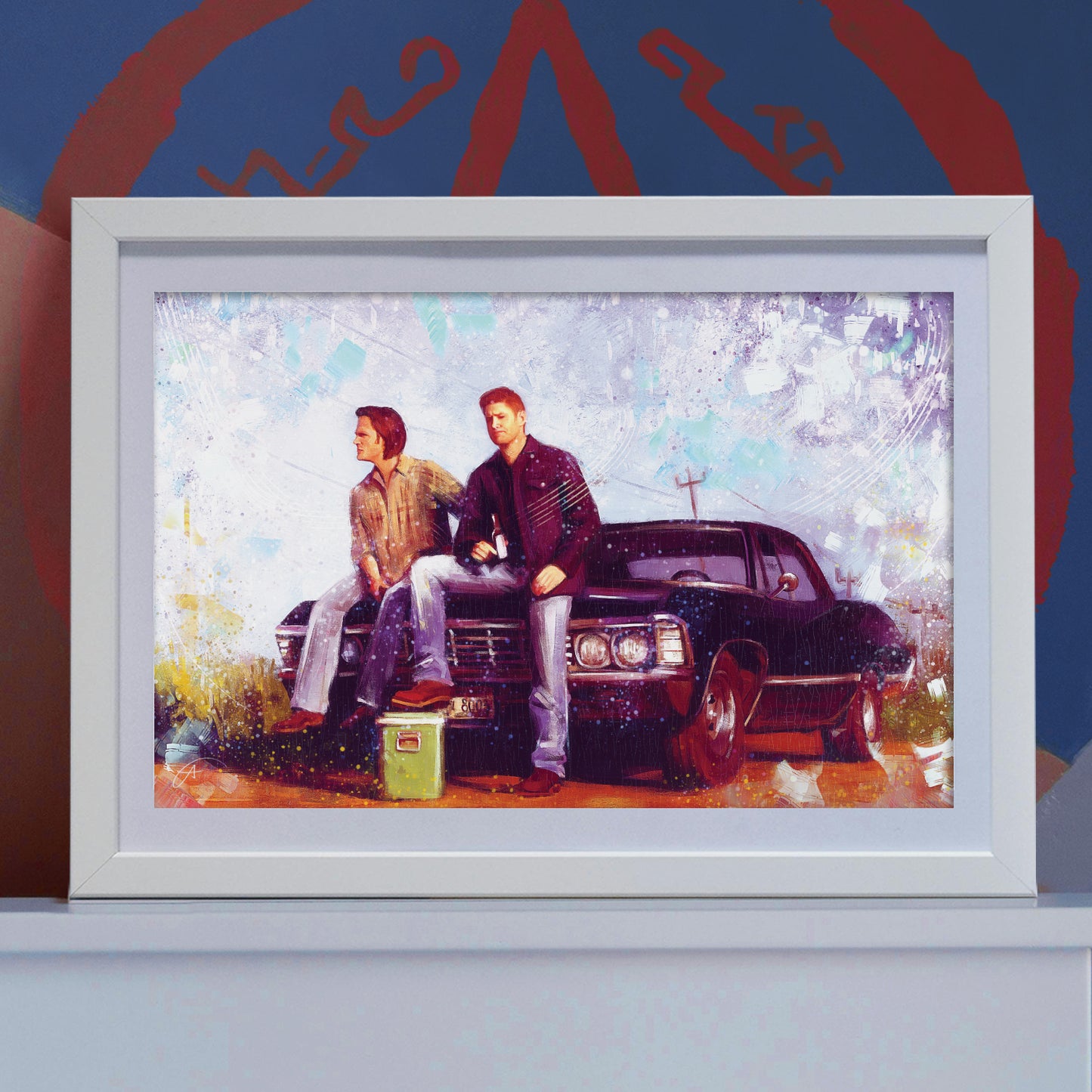 A picture in a white frame on a white shelf. The framed image shows Sam and Dean Winchester sitting on the hood of Baby, with a green cooler in front of the car. Behind the picture is a blue wall, with part of a Devil's Trap in red visible.
