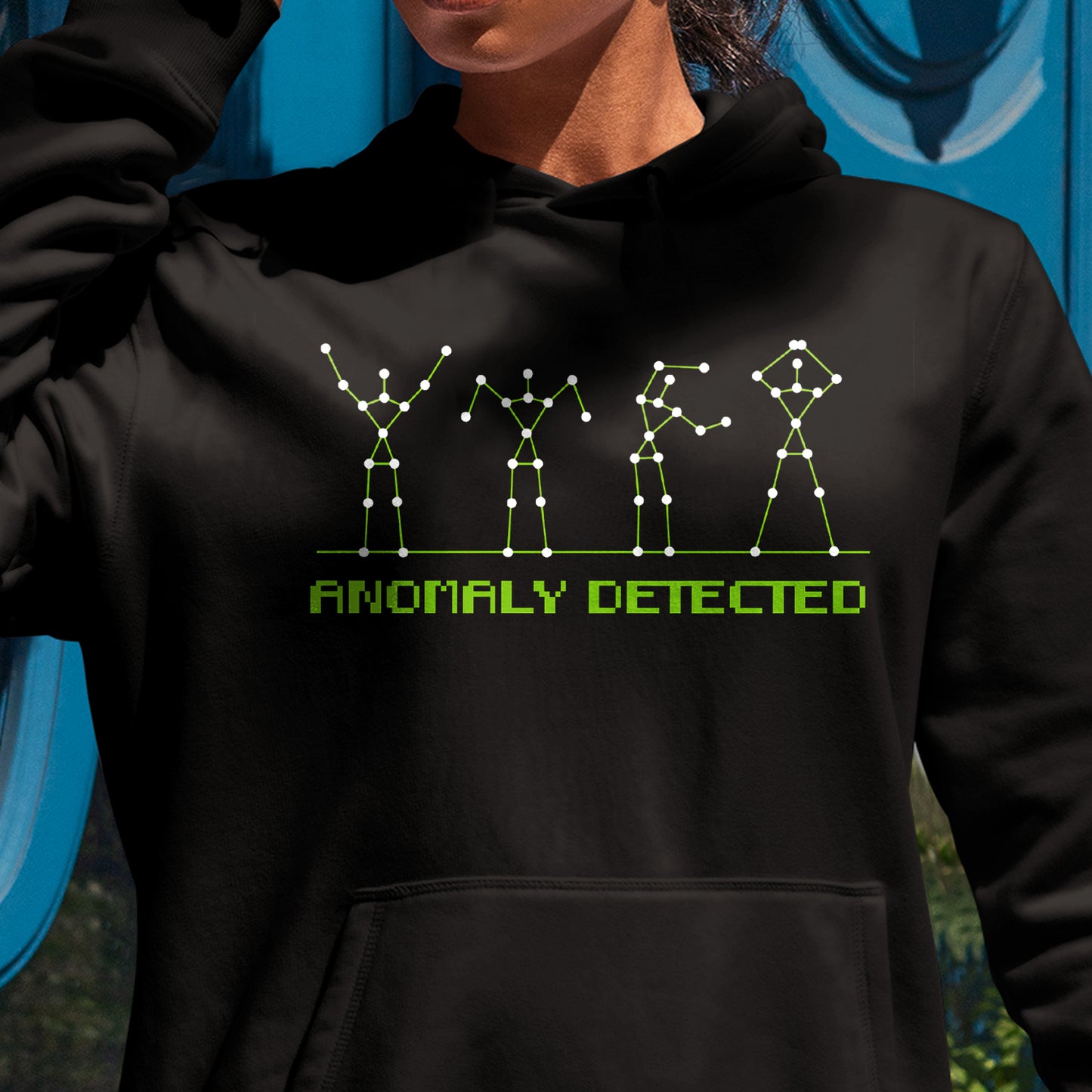 A female model in a black pullover hoodie. It reads "anomaly detected" in green 'digital' text. Above it are 4 figures standing on a thing green line. The figures are made out of thin green lines with white dot markers meant to resemble a chart reading, but they're forming the shapes of the 'YMCA' dance.