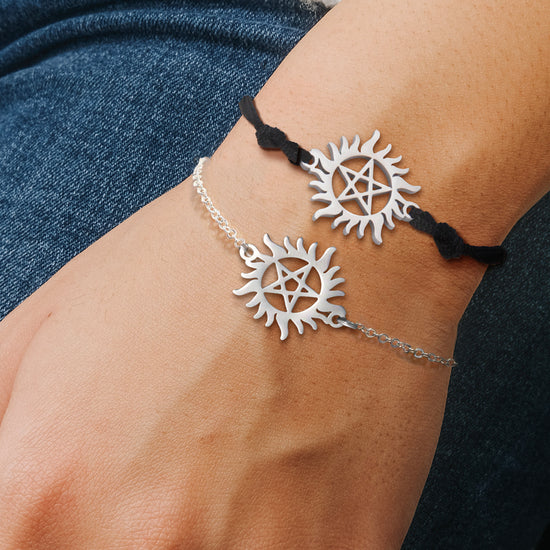 Close up of two bracelets on a model's wrist. Each bracelet has a charm in the center in the shape of the Anti-Possession symbol. The top bracelet has a stainless steel link chain attached to it. The bottom has a black braided cord attached to it.