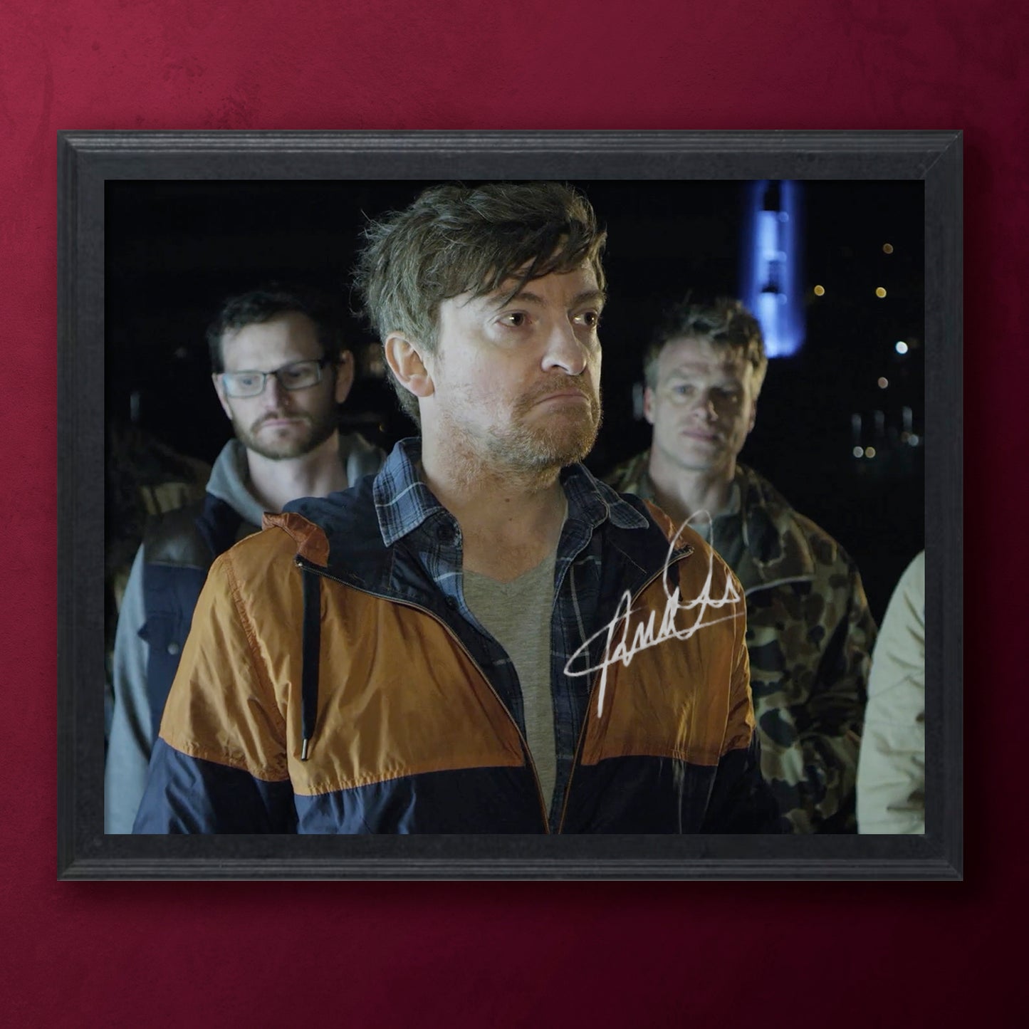 Framed photo of Rhys Darby with a serious expression, surrounded by others, on a red background.