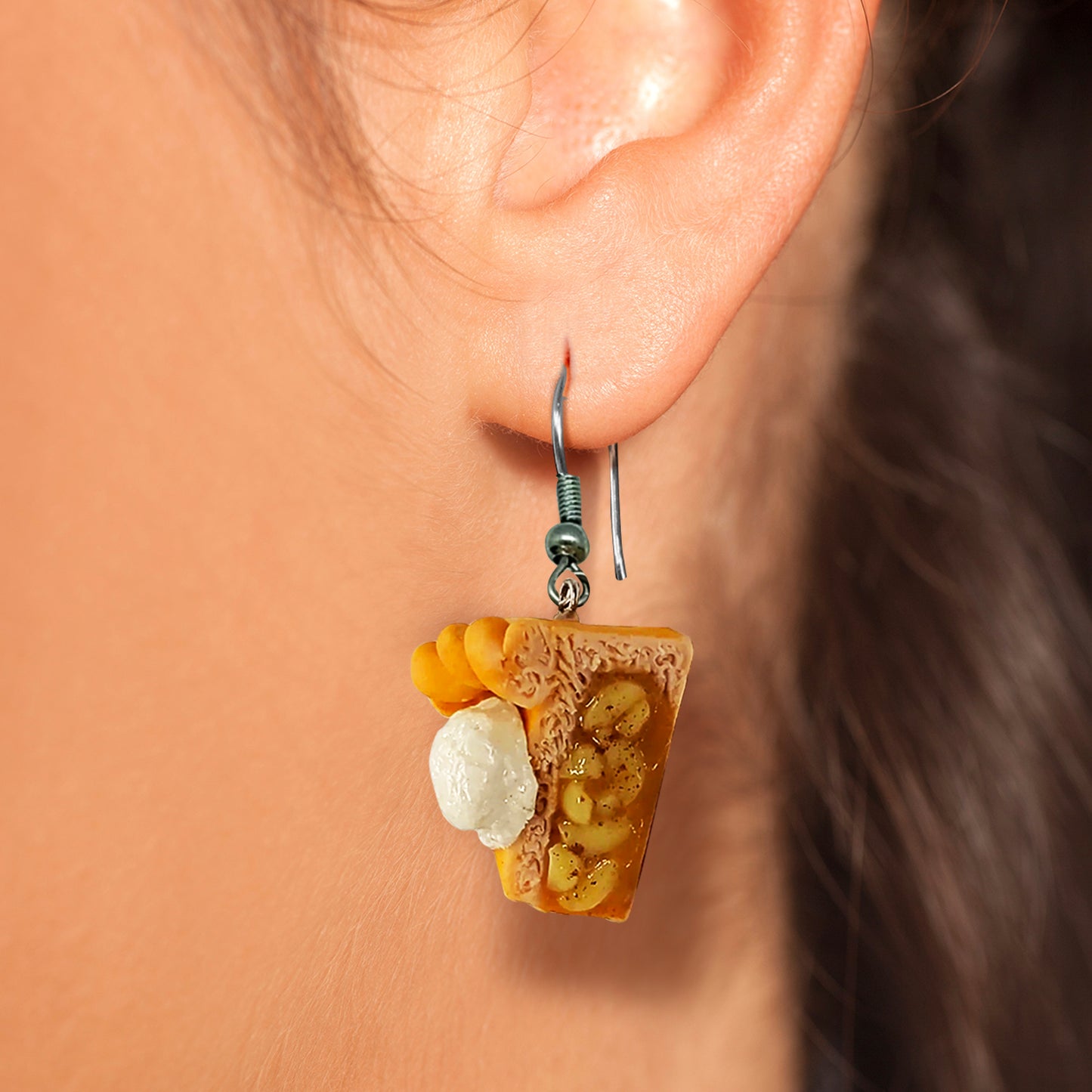 A close-up of a woman's ear. She is wearing a pair of small, clay earrings in the shape of apple pie slices. You can see the "apple filling" from the side of the earring, and there is a scoop of ice cream on top. The earrings are on a set of silver earring hooks.