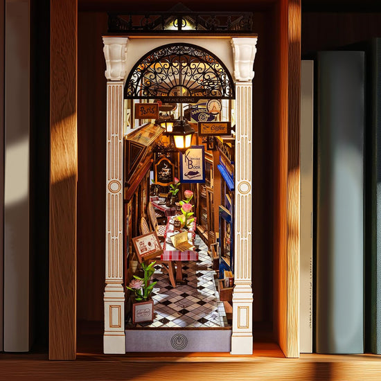 A miniature model kit depicting a covered arcade in Paris, on a shelf between books. The interior of the model has tables, shop fronts, and other details.