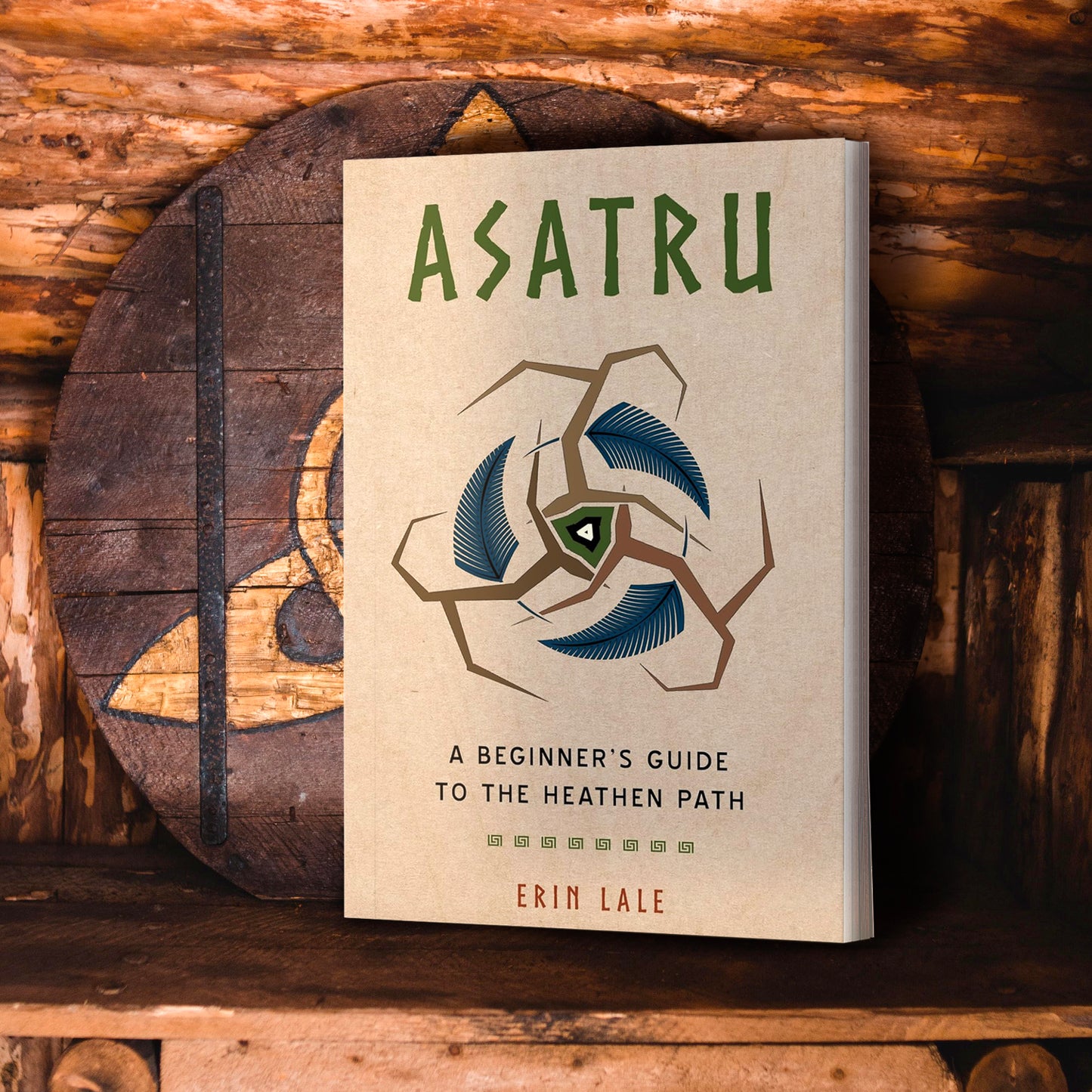A light brown book on a wooden shelf.. At the top of the cover is green text that says Asatru. Below the title is a brown symbol reselbling three tree branches, with a blue feather inside each branch. The center of the symbol is greenm white, and black. Under the symbol is black text saying A beginner's guide to the heathen path. Behind the book is a wooden shield with a norse rune.