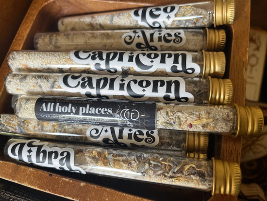 A collection of glass tubes with gold caps. Each tube has a sticker on the front, with black and white text listing the various names of the astrological signs. The tubes are sitting in a wooden box.