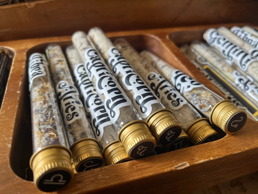 A collection of glass tubes with gold caps. Each tube has a sticker on the front, with black and white text listing the various names of the astrological signs. The tubes are sitting in a wooden box.