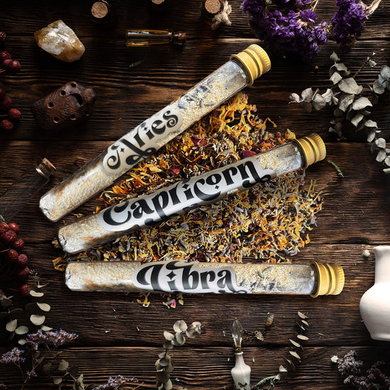 Three glass tubes with gold caps, lying on a wooden table surrounded by herbs, flowers, and crystals. Inside the tubes are bath salts and dried flowers. Each tube has a white label with the name of an astrological sign in black text.