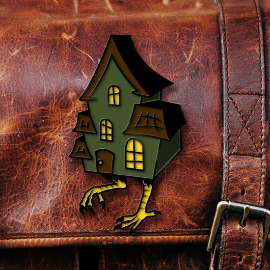 A green enamel pin on a brown leather bag. The pin is shaped like a multi-storied house, with yellow windows and brown roof sections. At the bottom of the house are two yellow chicken legs, in mid-step.