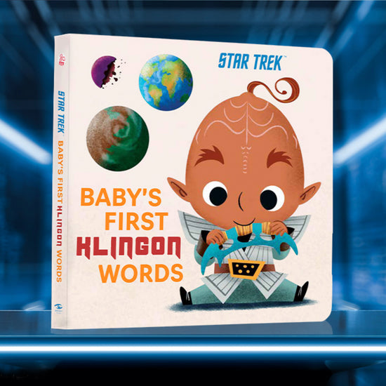 A book a futuristic blue table. On the cover is a cartoonish drawing of a baby Klingon from the Star Trek universe. The baby is wearing a Klingon warrior uniform, and chewing on a curved Klingon weaspon. Next to the baby are drawings of Earth and the Klingon homeworld. Orange and red text says "Baby's first klingon words." Behind the book is a blue hallway with long white lights.