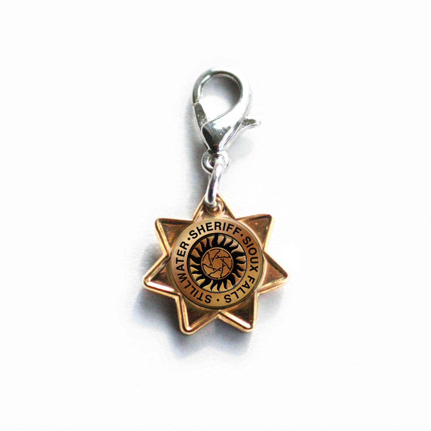 A brass charm in the shape of a Sheriff's badge. On the badge is a small anti-possession symbol encircled by the words SHERIFF - SIOUX FALLS - STILLWATER. The charm is on a silver bracelet/keychain clip. 