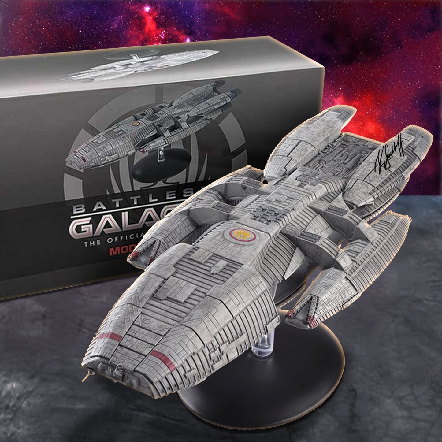 A detailed Battleship Galactica model spacecraft on a display stand, positioned in front of its branded product box. The model is signed in black ink and set against a dark, textured background with a red and purple nebula graphic.