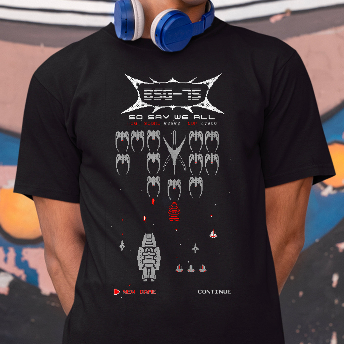 A person wearing a black graphic T-shirt with a retro video game style print featuring white and red elements. Blue headphones are around the neck, and the background is an outdoor wall with some colorful elements.