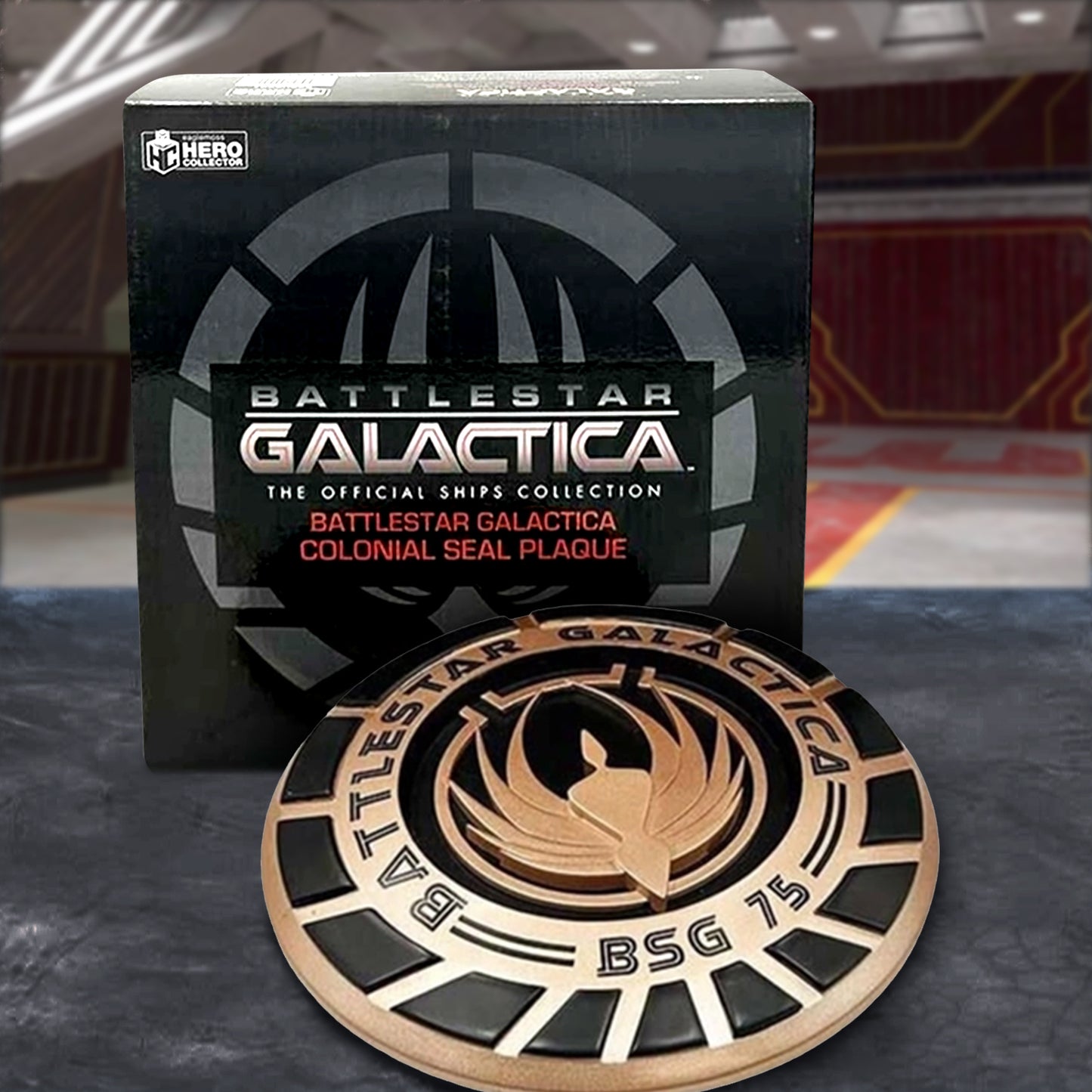 A bronze and black, round plaque, bearing the seal of the Battlestar Galactica. Behind the seal is a black box. The plaque and box are sitting on the floor of a launch bay.