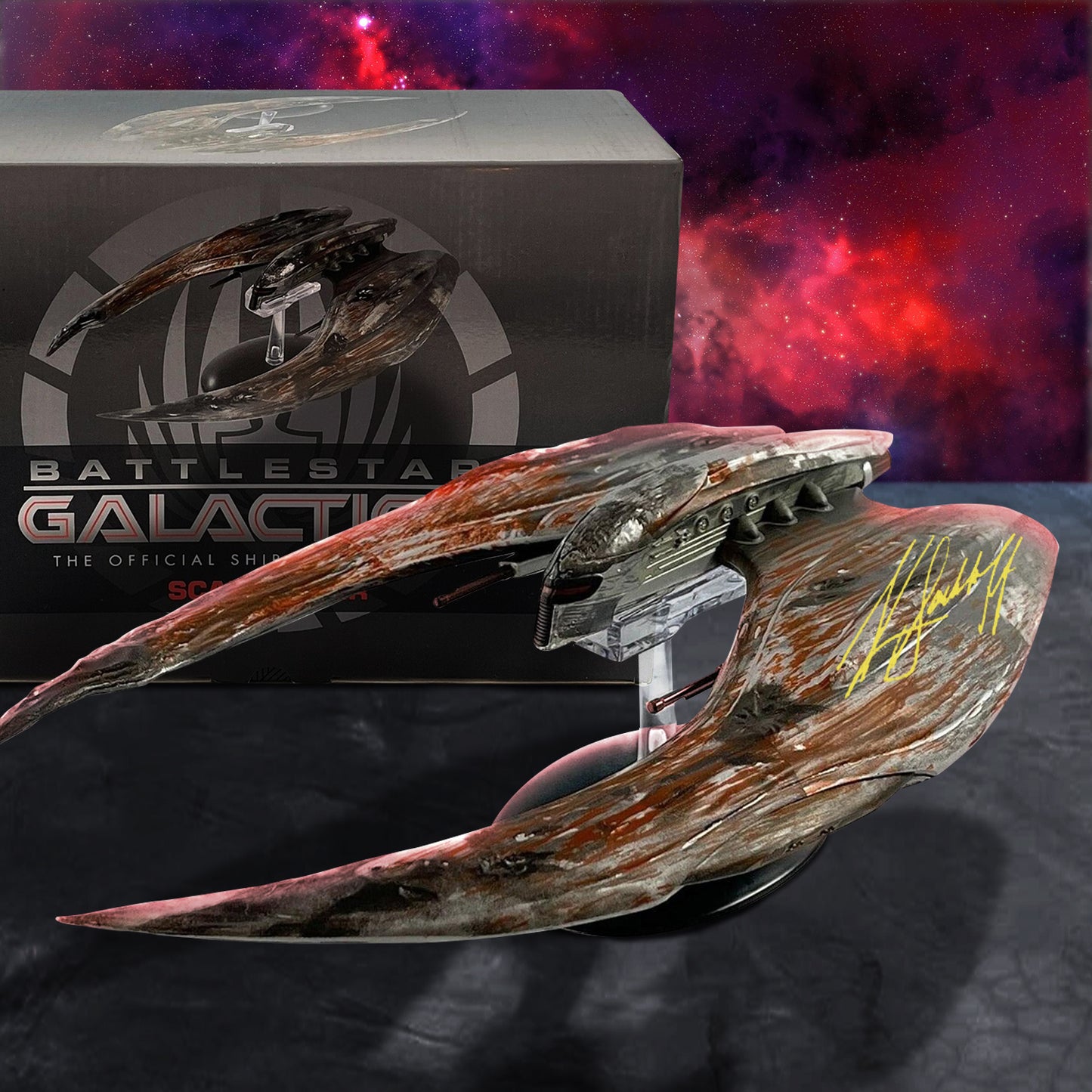 Battlestar Galactica official collectible model on a display stand, shown in front of its branded box against a red and purple nebula background. The model is signed in black marker on the outer wing.