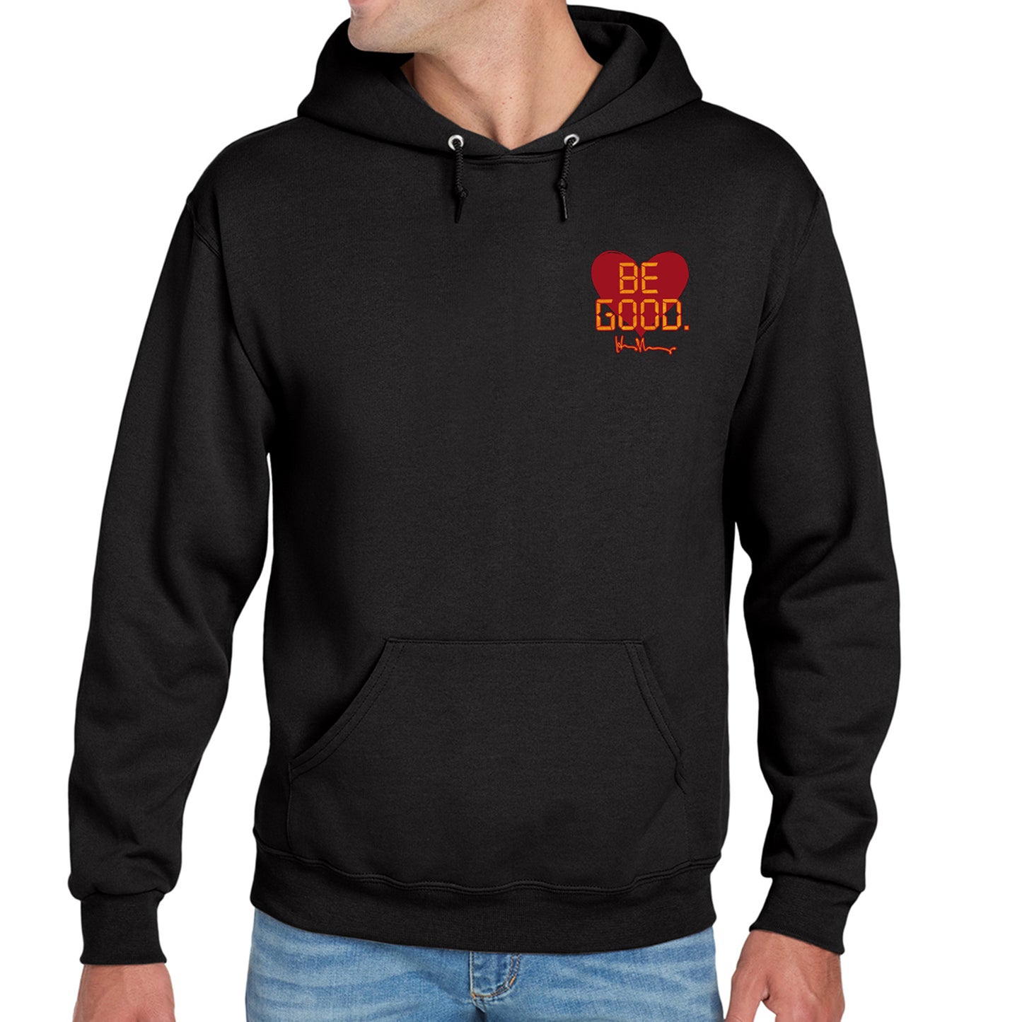 A model in a black hoodie against a white background. On the top left of the hoodie is a red heart. On top of the heart is "Be Good" in yellow text, stylized to look like text from old electronics.