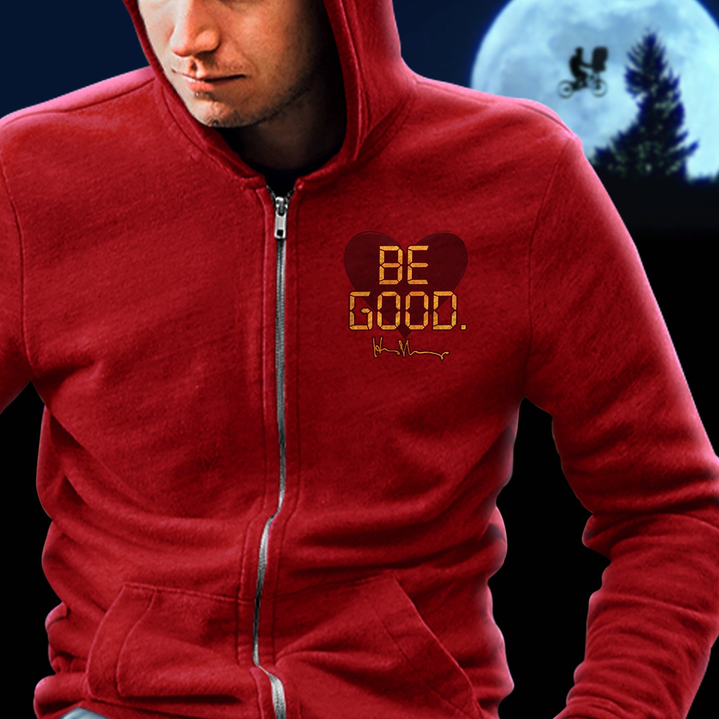 A model in a red zip hoodie against a black background. On the top left of the shirt is a red heart. On top of the heart is "Be Good" in yellow text, stylized to look like text from old electronics. Behind the model is a full moon with a silhouette of a kid on a bike flying in front of it.