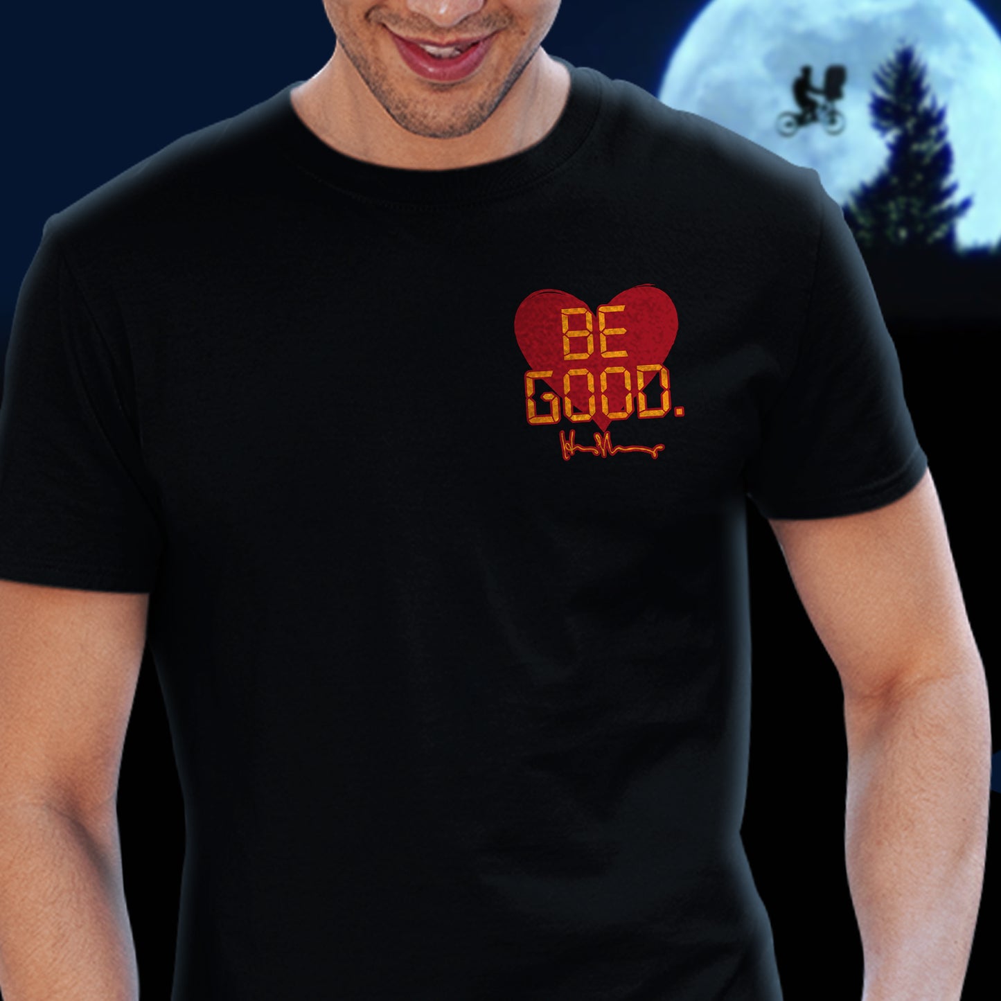A model in a black tee shirt against a black background. On the top left of the shirt is a red heart. On top of the heart is "Be Good" in yellow text, stylized to look like text from old electronics. Behind the model is a full moon with a silhouette of a kid on a bike flying in front of it.