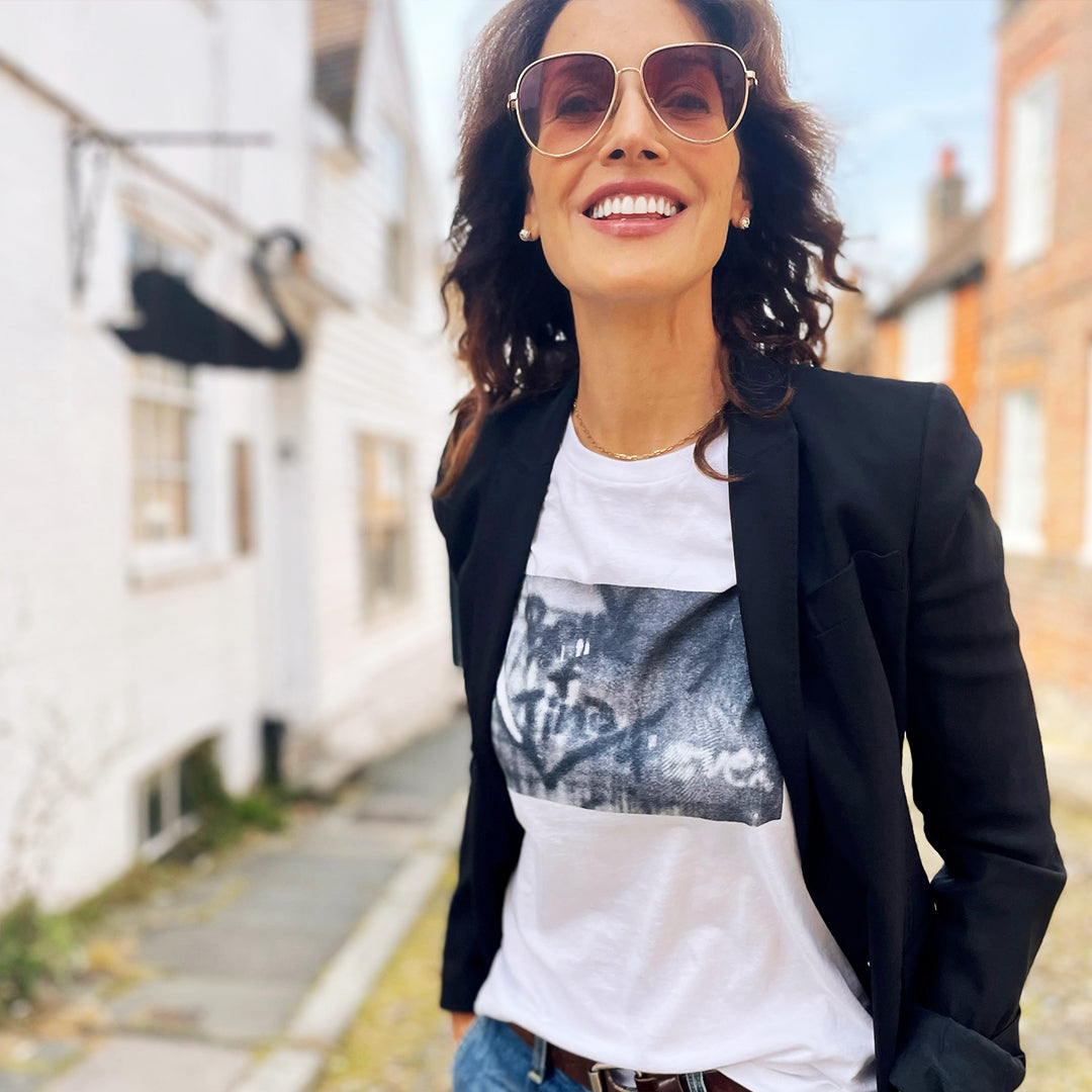 An image of actress Jennifer Beals in a white t-shirt. and black blazer. On the shirt is a black and white wall with Bette + Tina 4 Ever spray painted inside a heart. Behind Jennifer is an alley between houses.