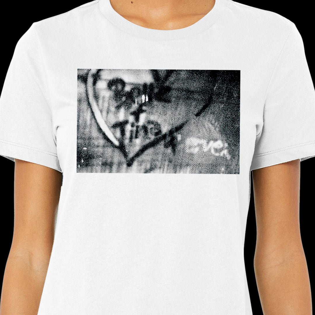A model wearing a white T-shirt, against a black background. On the shirt is a black and white wall with Bette + Tina 4 Ever spray painted inside a heart.