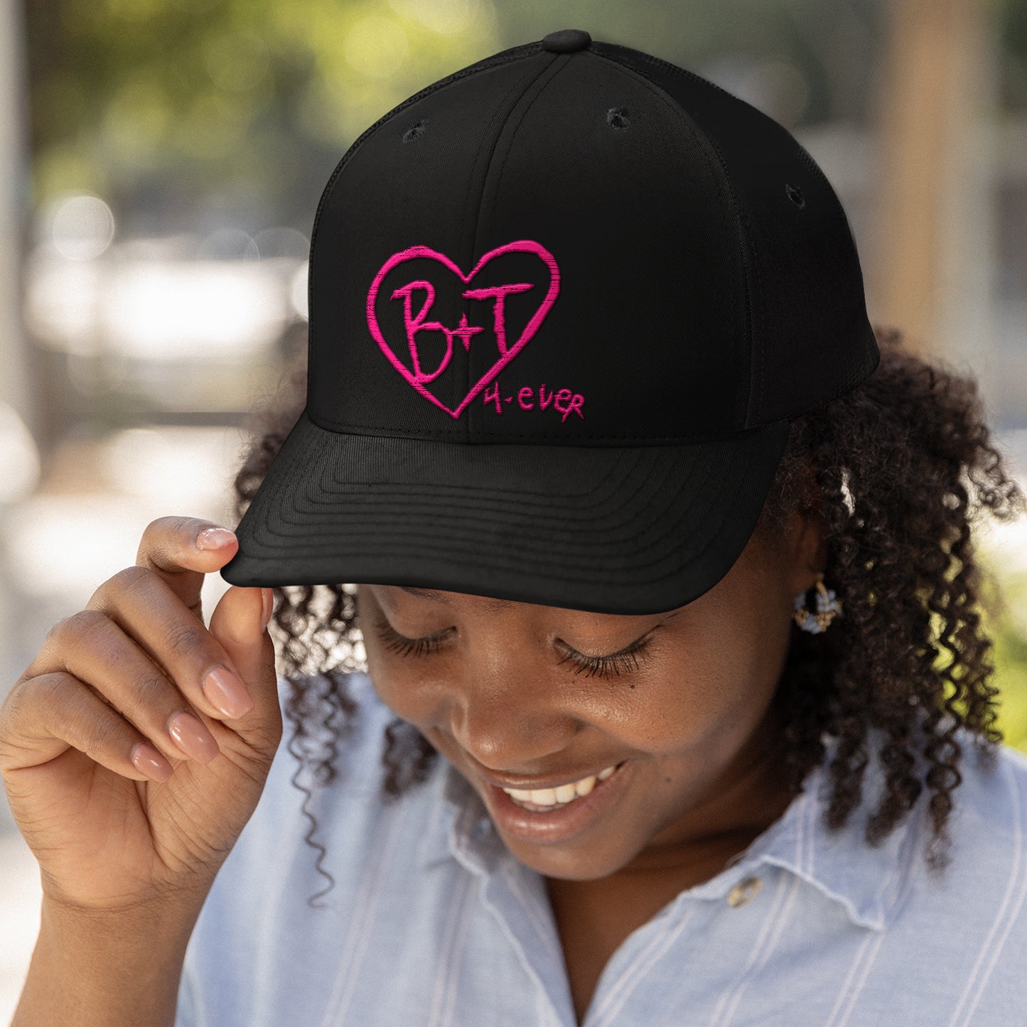 A female model wearing a black baseball hat. On the front of the hat is a pink heart with B & J inside it. Next to the heart is pink text saying 4 ever. Behind the model is a blurred park area.