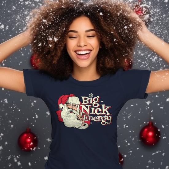 A woman with dark skin and curly hair wearing a navy-blue unisex tee. The tee features the words "Big Nick Energy" in a vintage, cream-and-white font, alongside an illustration of Santa Claus.