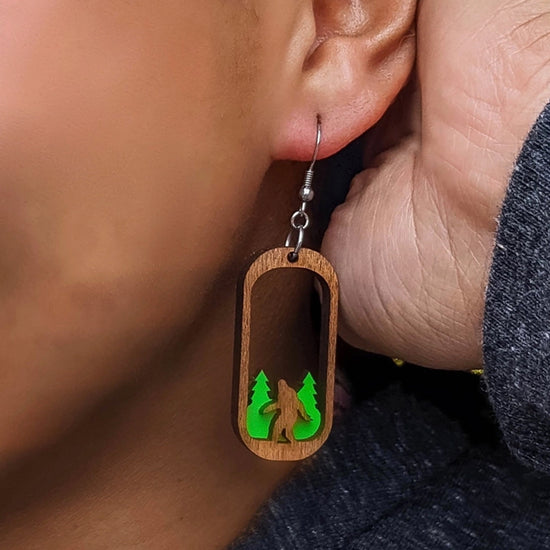 Close up view of a model wearing oval-shaped wooden earrings, with the center of each cut away. At the bottom of each earring is a carved Bigfoot figure, standing in front of green trees.