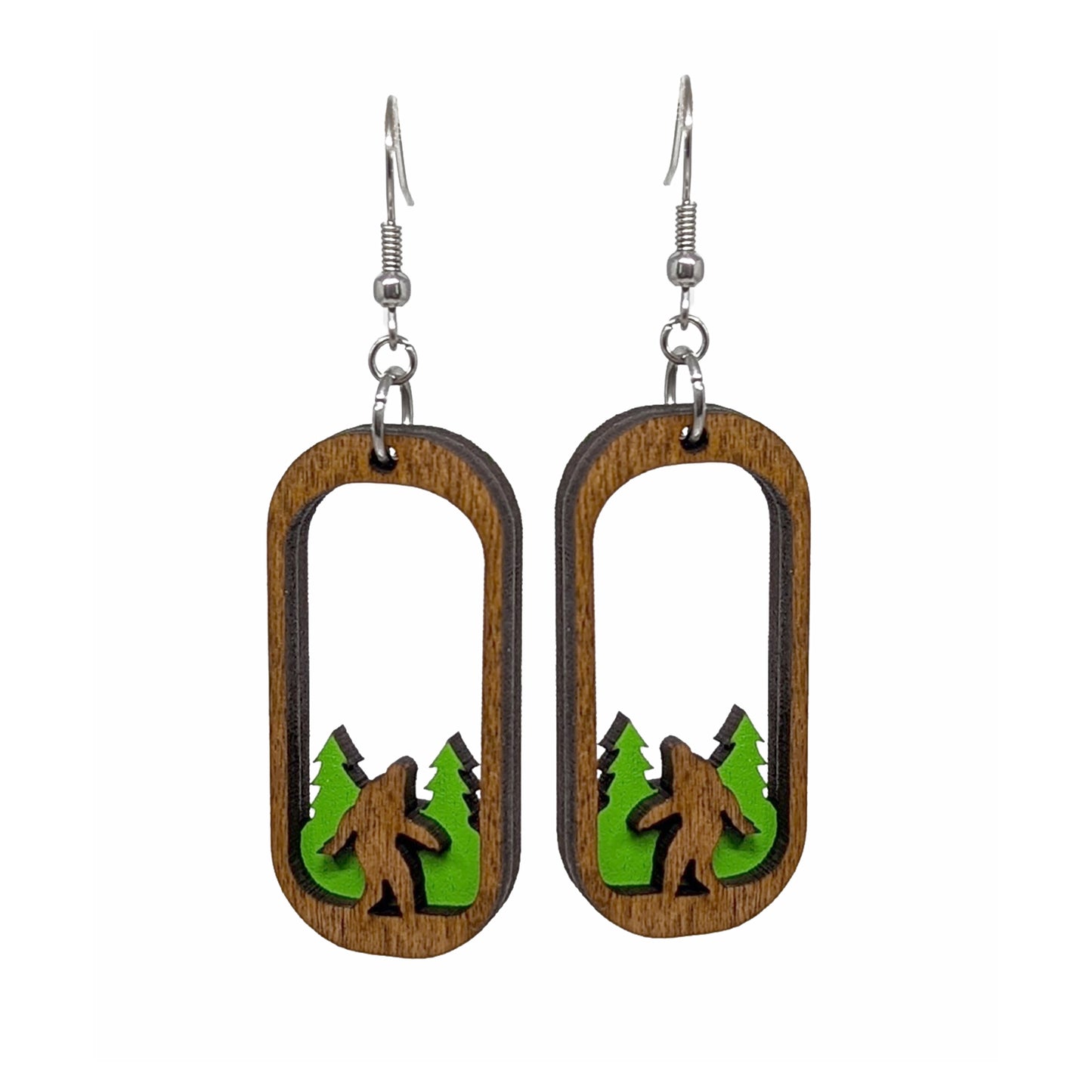 Load image into Gallery viewer, Two oval-shaped wooden earrings on a white background, with the center of each cut away. At the bottom of each earring is a carved Bigfoot figure, standing in front of green trees.
