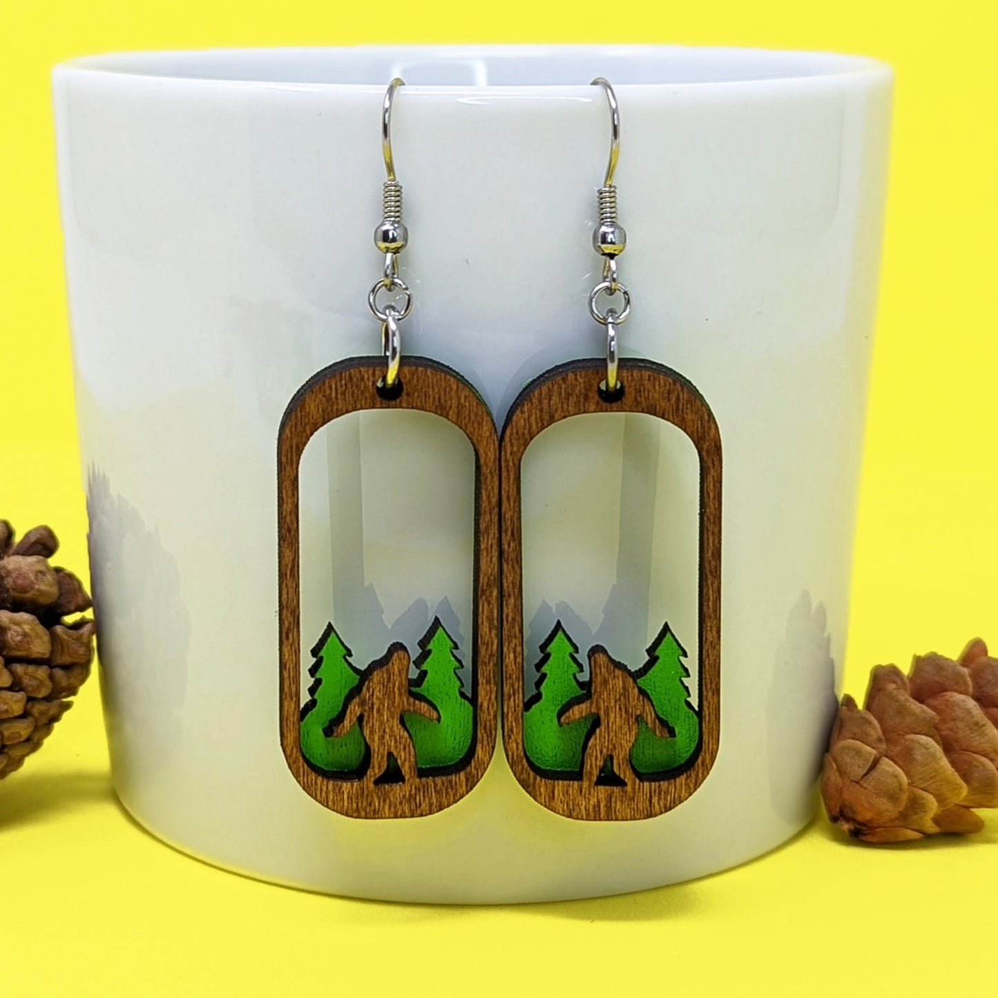 Load image into Gallery viewer, A white mug on a yellow background with pinecones on either side. Two oval-shaped wooden earrings hang from the mug, with the center of each cut away. At the bottom of each earring is a carved Bigfoot figure, standing in front of green trees.
