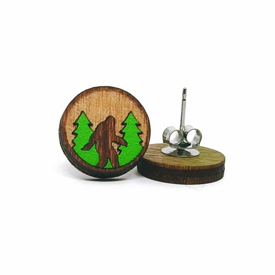 A round wood earring on a white background. In the center is a carved Bigfoot figure, standing in front of green trees. A second earring is next to it, turned upside down to show the post.